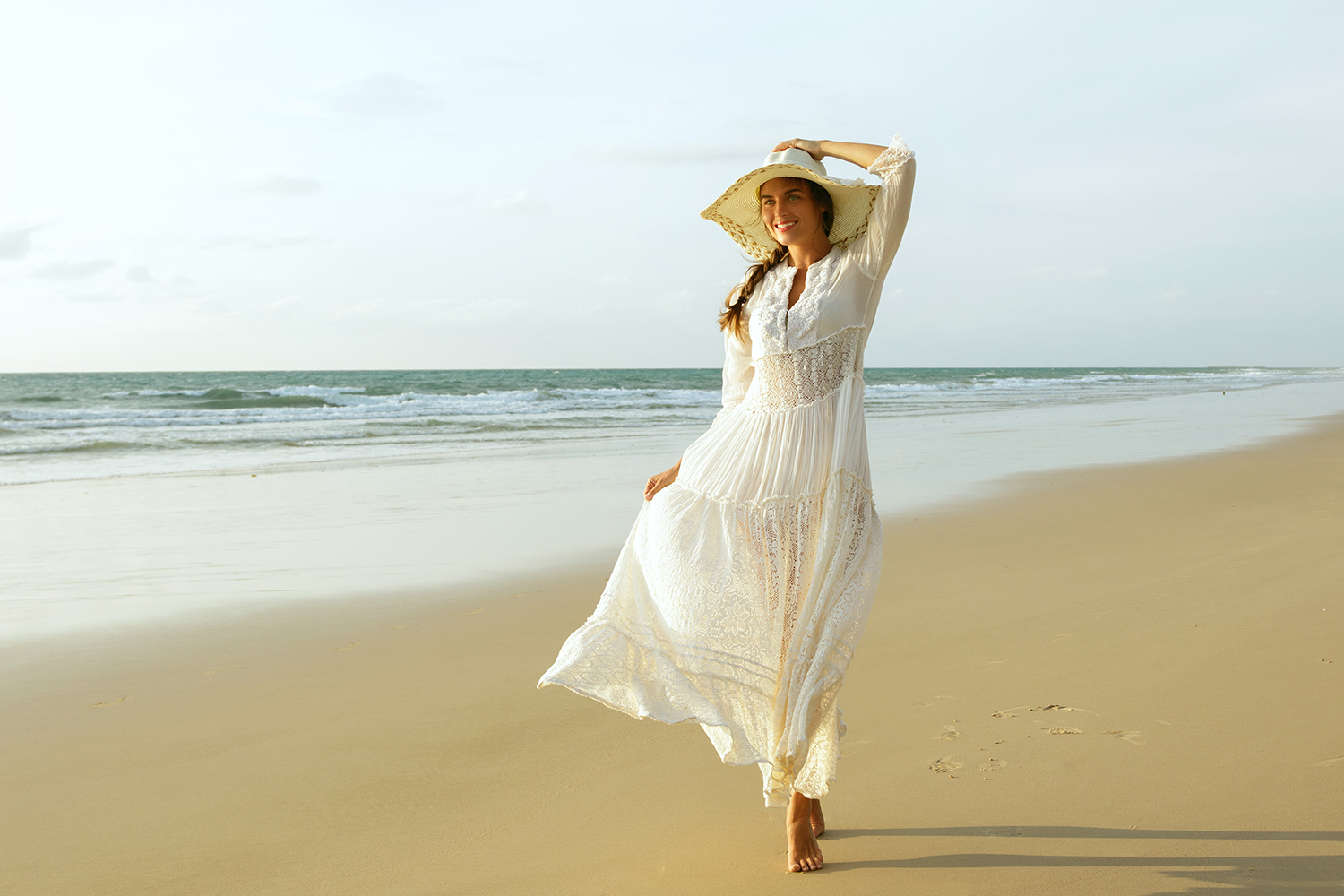 Happy young woman wearing beautiful white dress is walking on the beach during sunset
