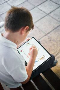 One little spanish school boy drawing on a tablet sitting on bench in park